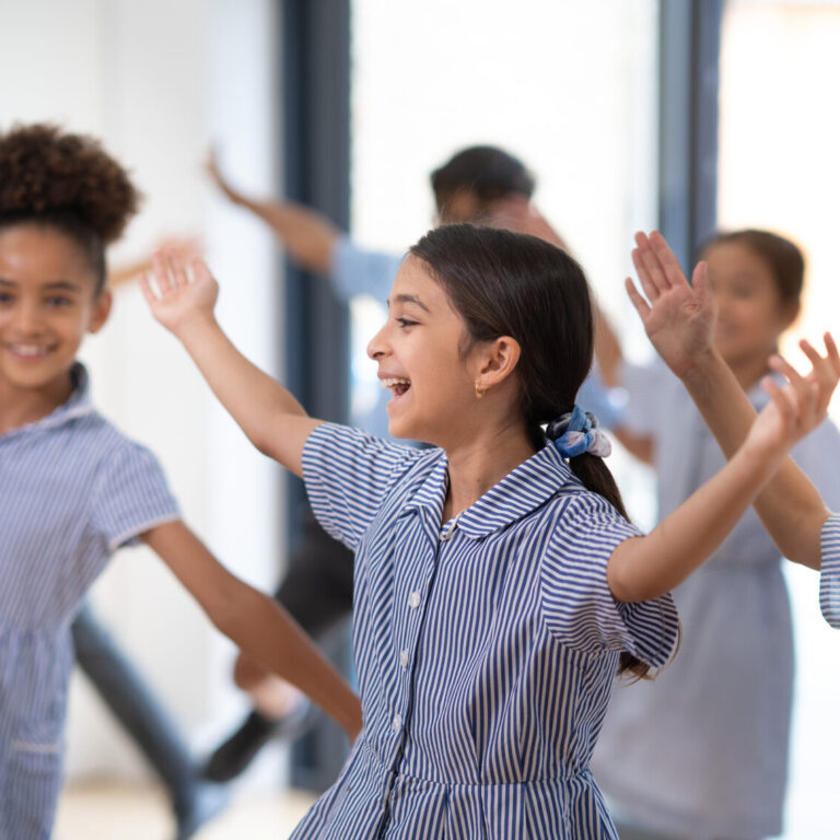 Group of children in blue striped dresses dancing with arms raised in a bright hallway, smiling and enjoying the activity