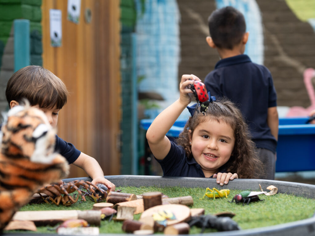 Smiling girl in a navy shirt raises a red spotted toy as she plays with wooden blocks in a sandbox-like area, other children and a fence in the background.