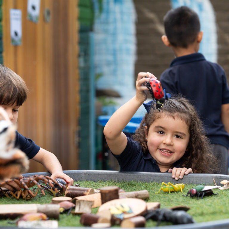 Smiling girl in a navy shirt raises a red spotted toy as she plays with wooden blocks in a sandbox-like area, other children and a fence in the background.