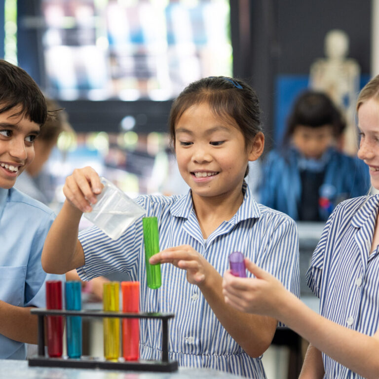 Three students in striped uniforms work at a science lab bench, handling colorful test tubes and a pink clipboard while smiling at each other.